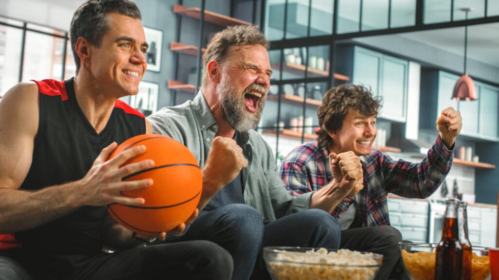 Three men cheer enthusiastically while watching a game on TV. One holds a basketball, and snacks and drinks are on the table in front of them.