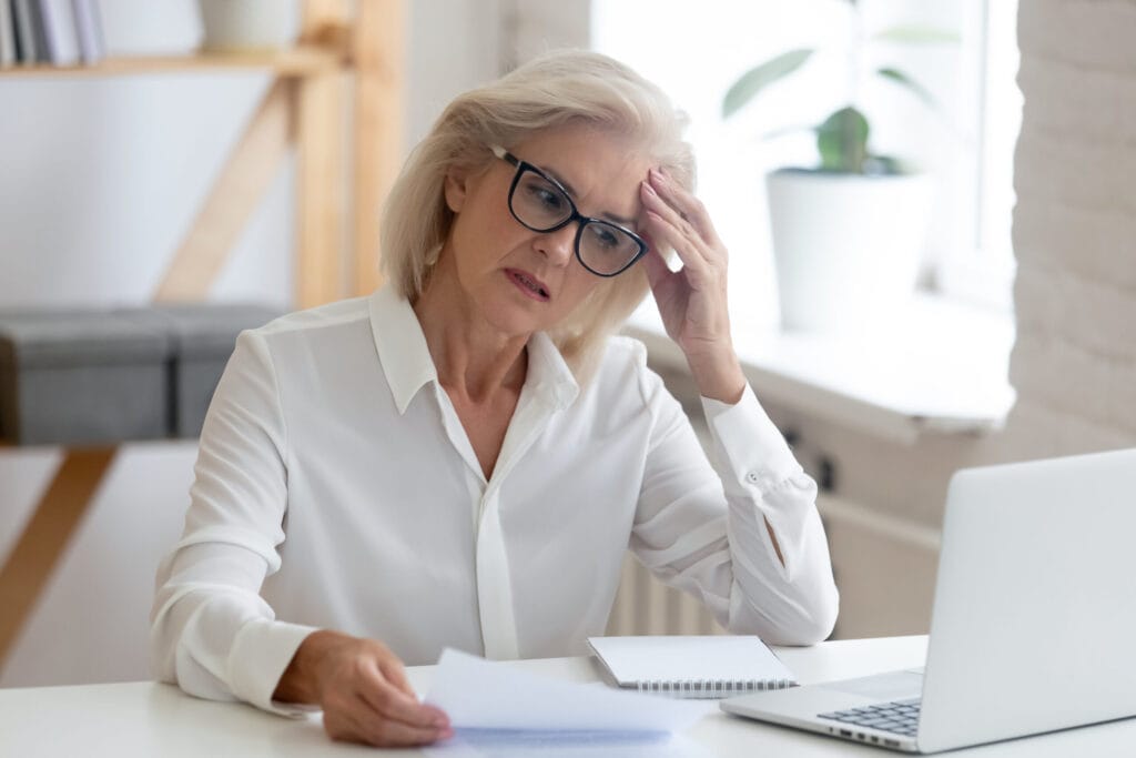 A woman wearing glasses, a white shirt, and sitting at a desk looks stressed while holding papers and looking at a laptop.