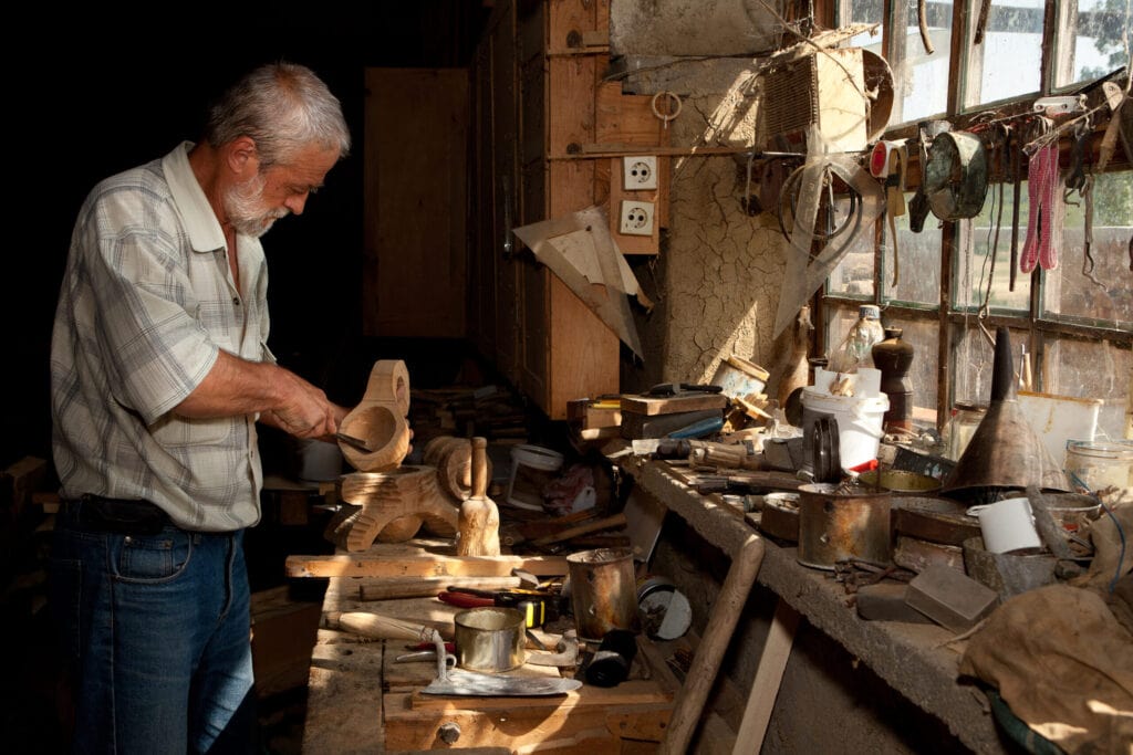 An older man with a gray beard works on a wooden object in a cluttered workshop with various tools and supplies on the bench by a window.