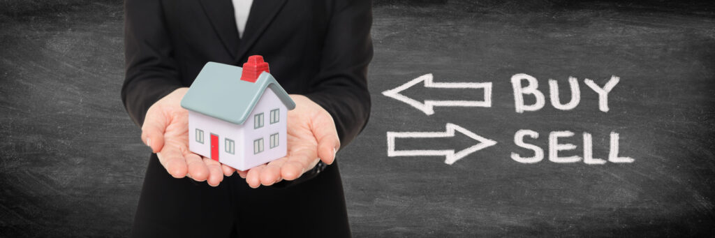 Person in a suit holding a small house model with arrows pointing to "BUY" and "SELL" written on a chalkboard in the background.