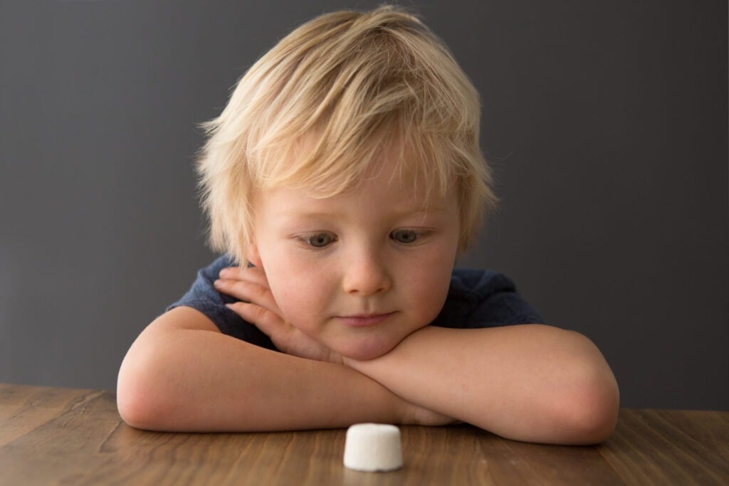 A young child with blonde hair, resting their head on their crossed arms, looks intently at a white marshmallow on a wooden table against a dark background.