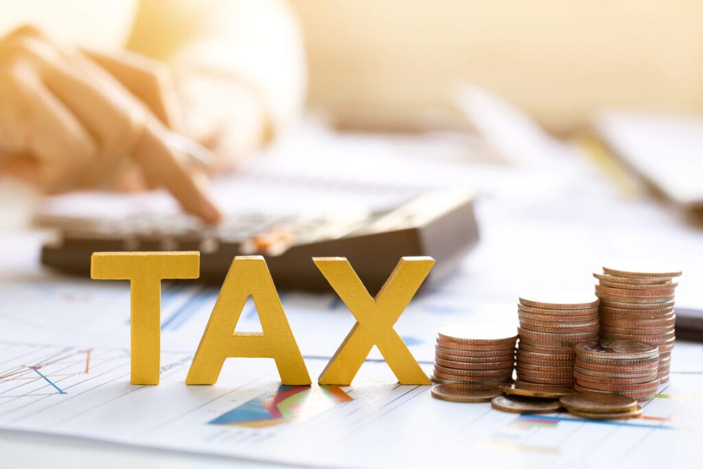 A person is using a calculator with the word "TAX" and stacks of coins on a desk filled with papers in the foreground.