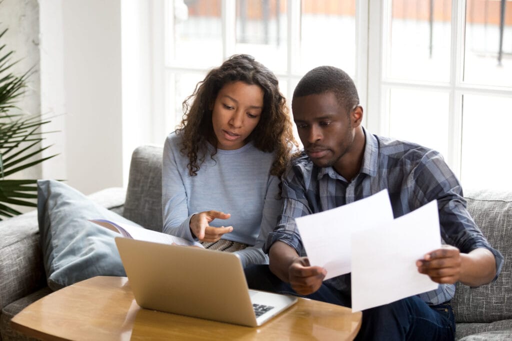 A woman and a man sit on a couch looking at documents and a laptop. The woman points at the laptop screen while the man holds papers.