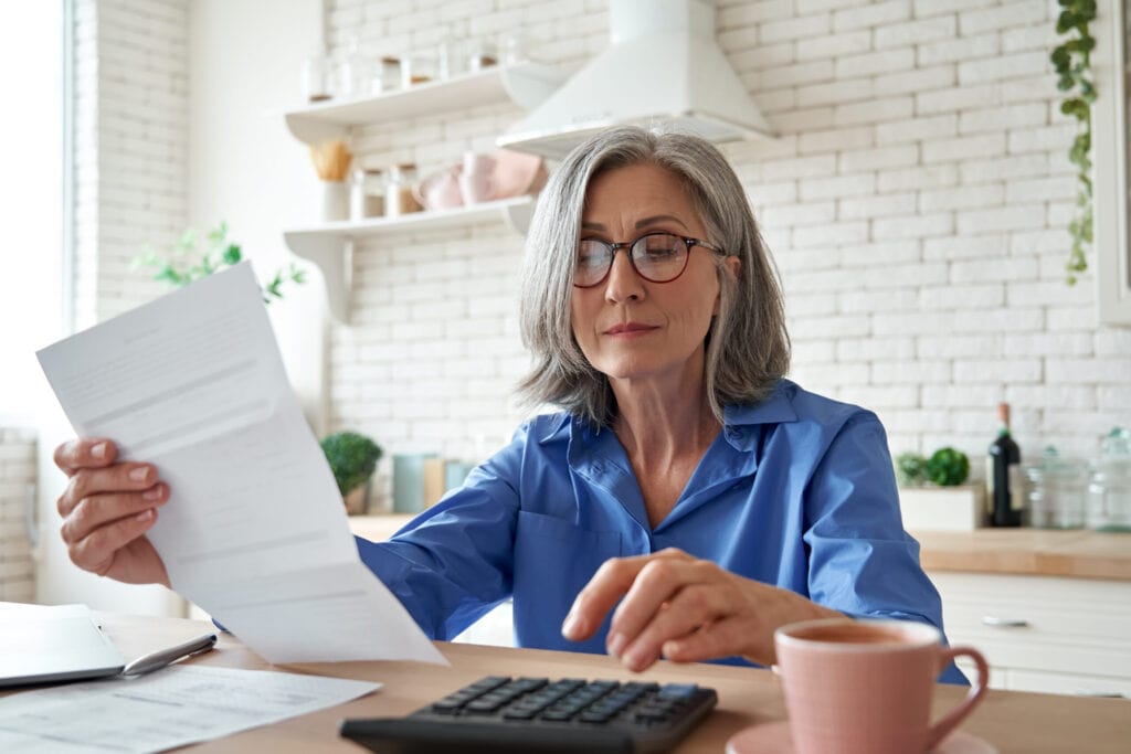 A woman with glasses, wearing a blue shirt, sits at a table in a kitchen, reading a document and using a calculator. A pink mug is on the table beside her.