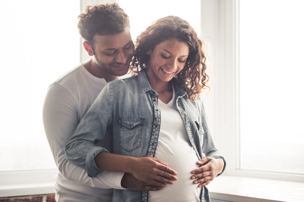 A couple stands together near a window; the man is embracing the woman from behind, both smiling and holding her pregnant belly.