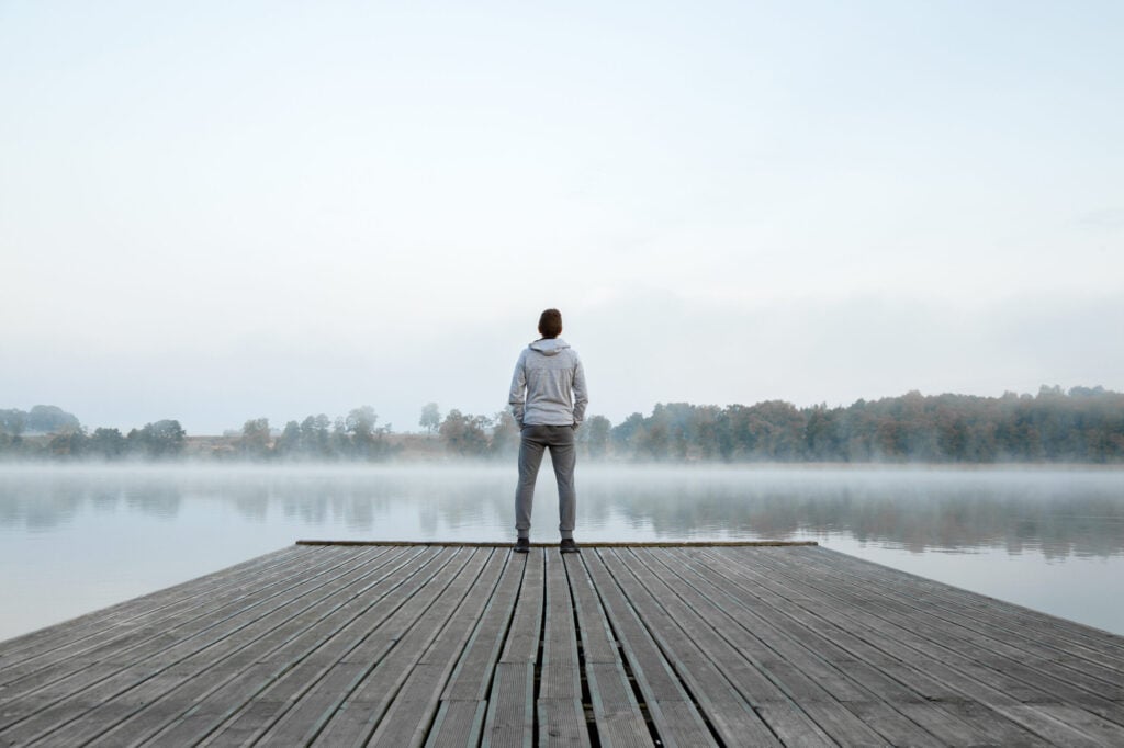 A person stands at the end of a wooden dock, looking out over a calm, foggy lake with trees in the background.