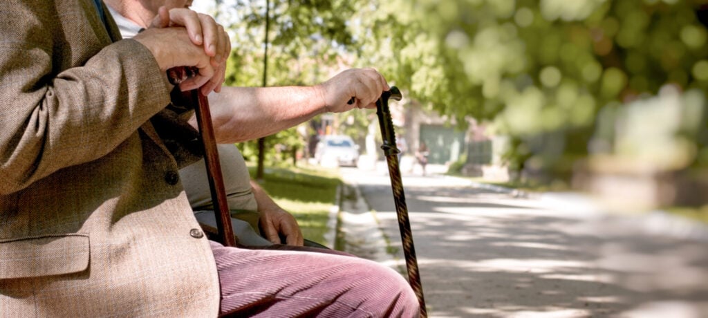 An elderly person wearing a blazer and holding a cane sits on a bench in a sunlit park, with greenery and a pathway in the background.
