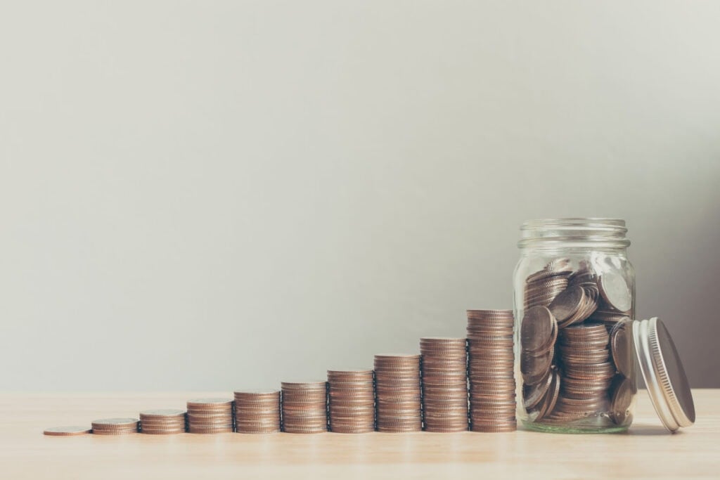 A row of stacked coins gradually increasing in height leads to an open glass jar filled with coins on a wooden surface against a plain background.