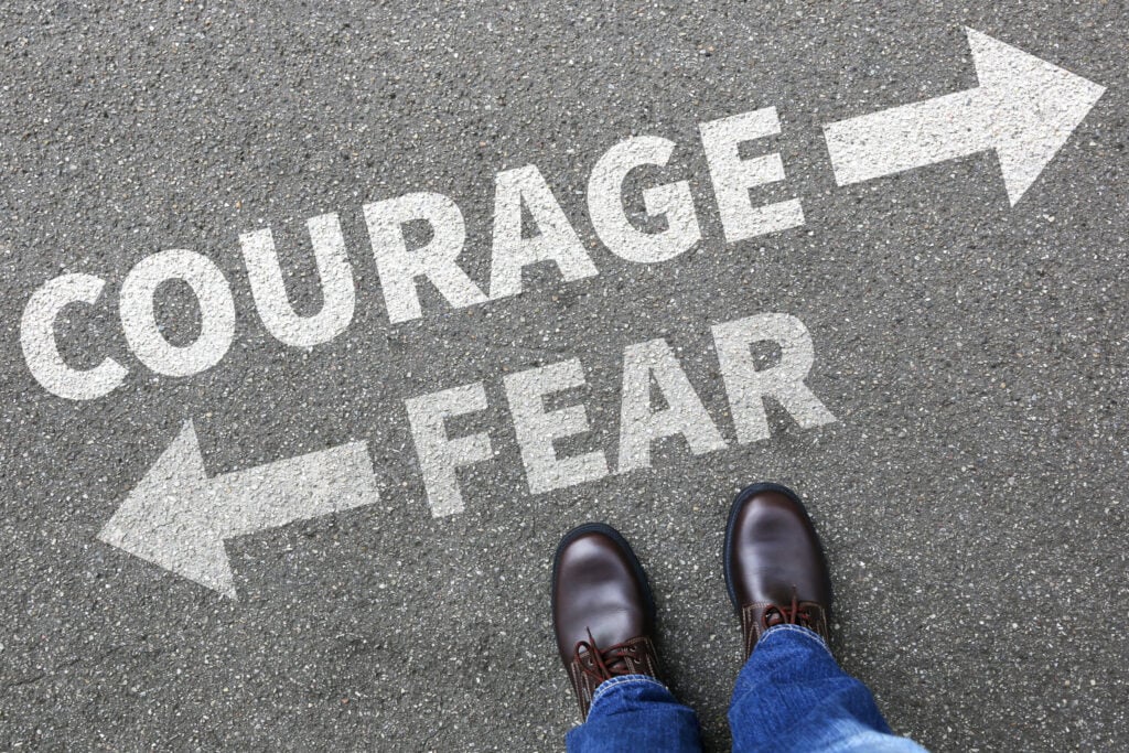 A pair of feet standing on a road with the words "Courage" and "Fear" written in white, each with an arrow pointing in opposite directions.