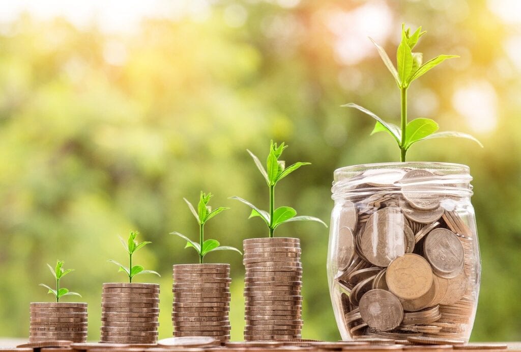 Four stacks of coins, each topped with a small plant, increase in size from left to right. On the far right is a jar filled with coins and topped with a larger plant. The background is blurred greenery.