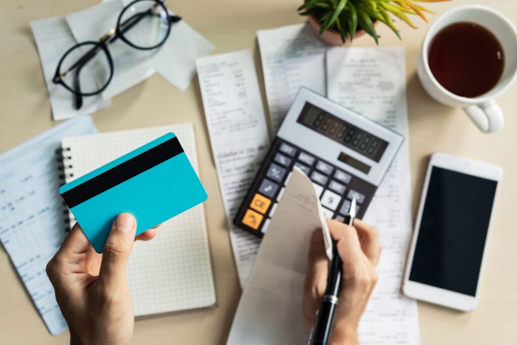 Person holding a blue credit card while reviewing financial documents, surrounded by a notebook, eyeglasses, calculator, smartphone, and cup of tea.