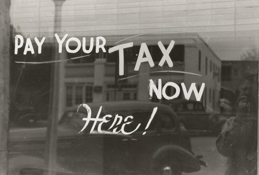 A vintage storefront window sign reads "Pay Your Tax Now Here!" against the reflection of a street scene with cars and buildings.