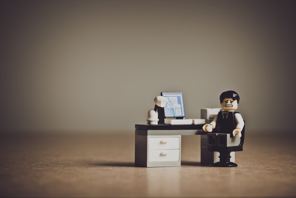 A Lego figurine dressed in business attire sits at a small black and white office desk with a computer and lamp on it, set against a plain background.