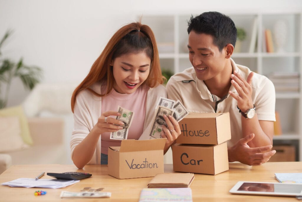 A couple sits at a table, smiling and inserting money into boxes labeled "Vacation," "House," and "Car." A calculator and papers are on the table beside them.