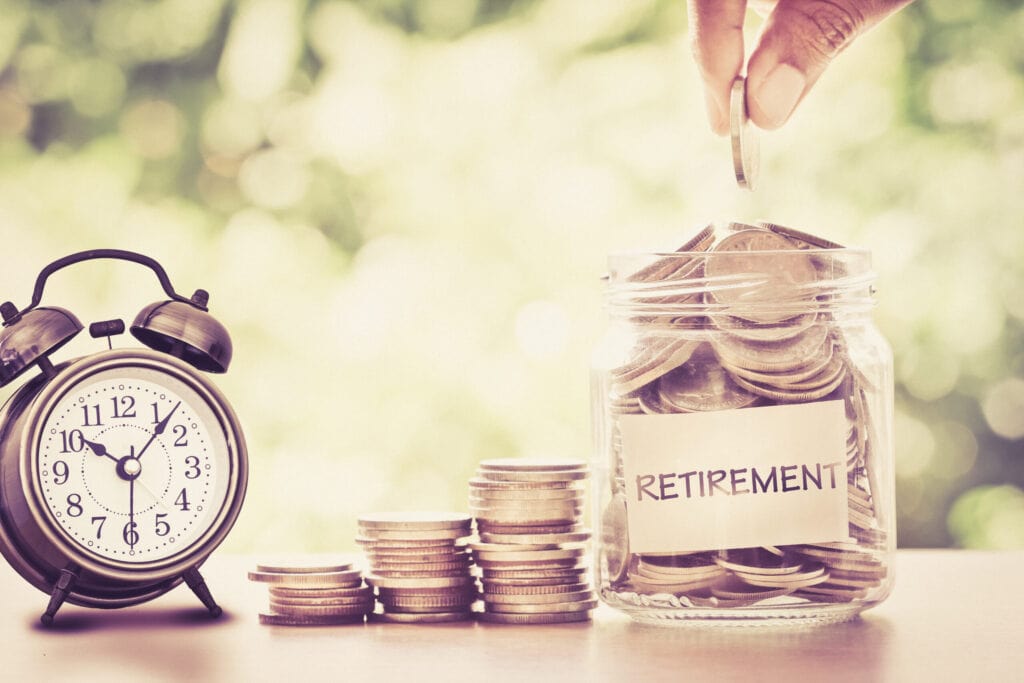 A hand drops a coin into a jar labeled "Retirement" filled with coins. An old-fashioned alarm clock and stacks of coins sit next to the jar on a table, with a blurred green background.