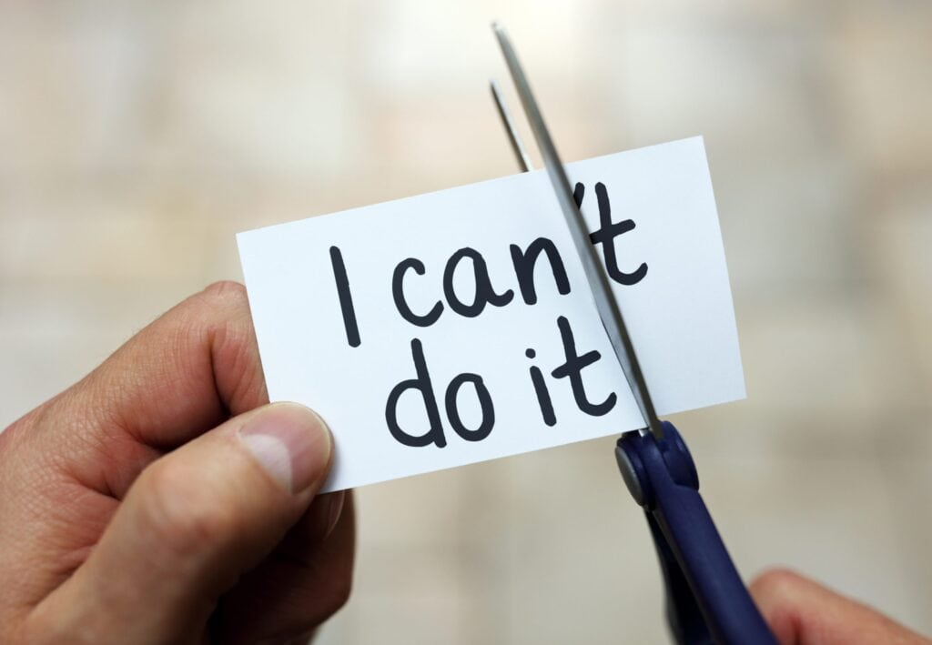 A hand is cutting a piece of paper using scissors. The paper initially says "I can't do it" but the person is cutting off the "t," changing the message to "I can do it.