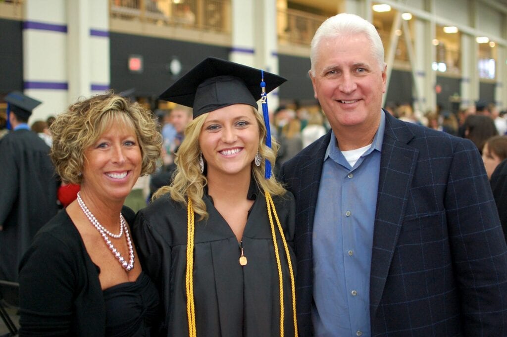 A young woman in graduation attire stands smiling between two adults, likely her parents, at an indoor event.
