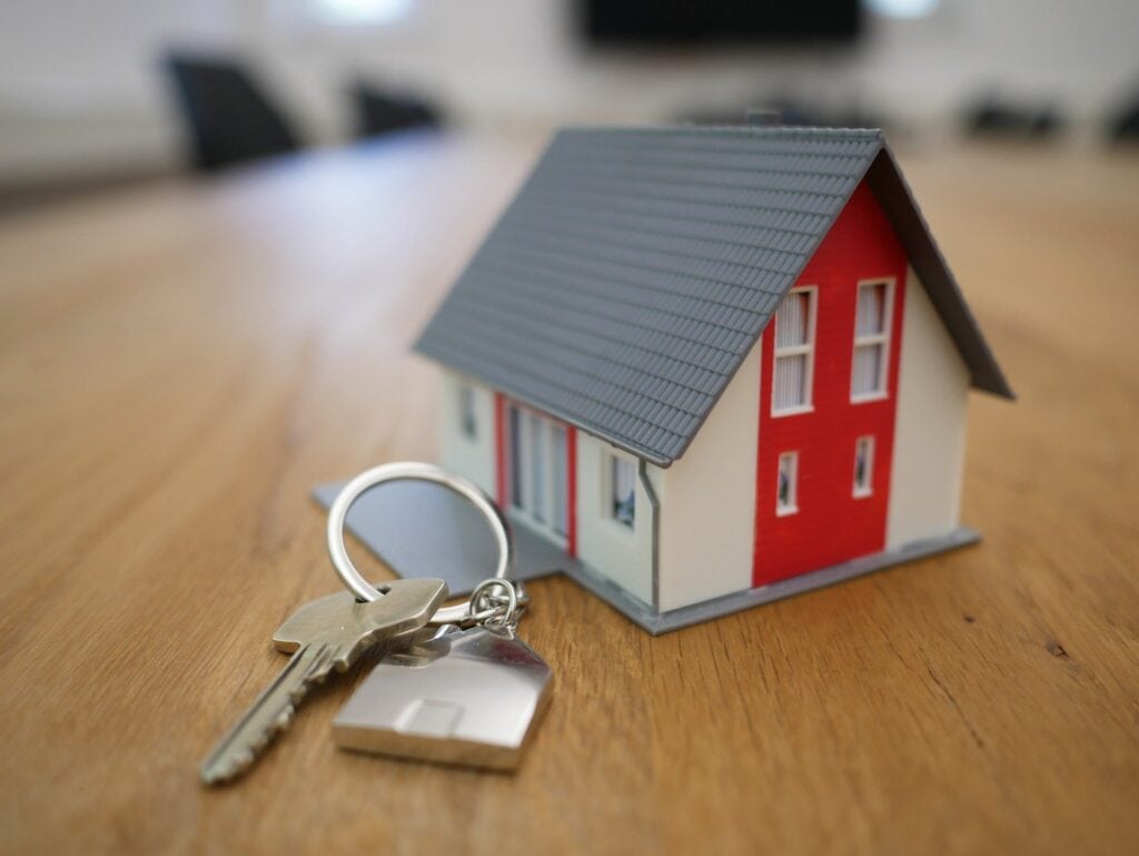 A small model house with a gray roof and red accents sits on a wooden surface next to a keychain with two keys.