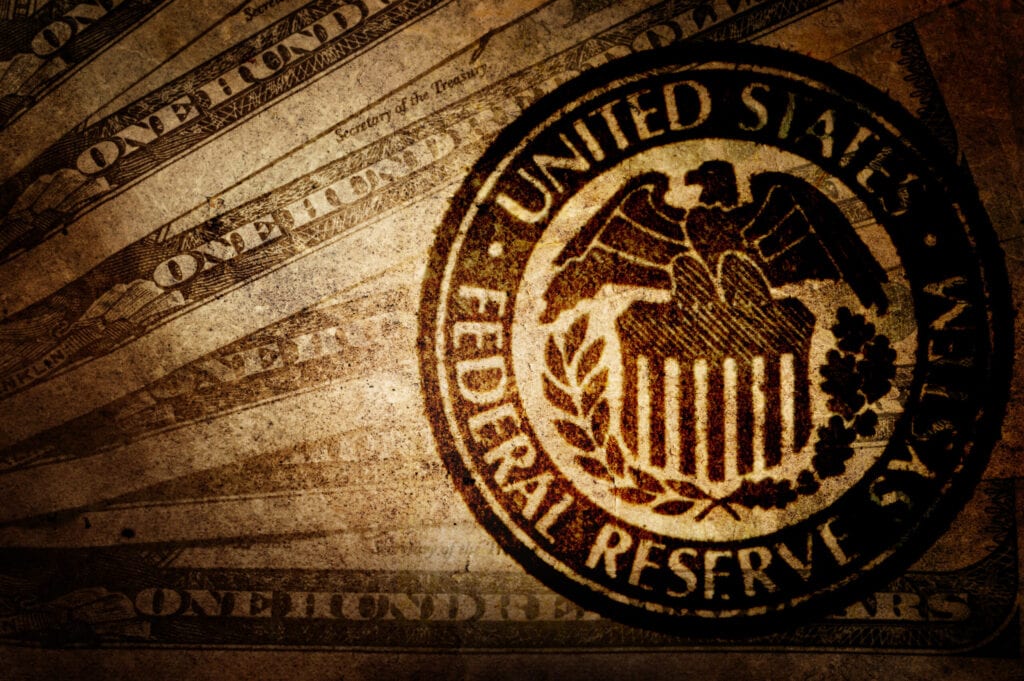 The image shows the seal of the United States Federal Reserve System overlaid on a background of various US dollar bills.