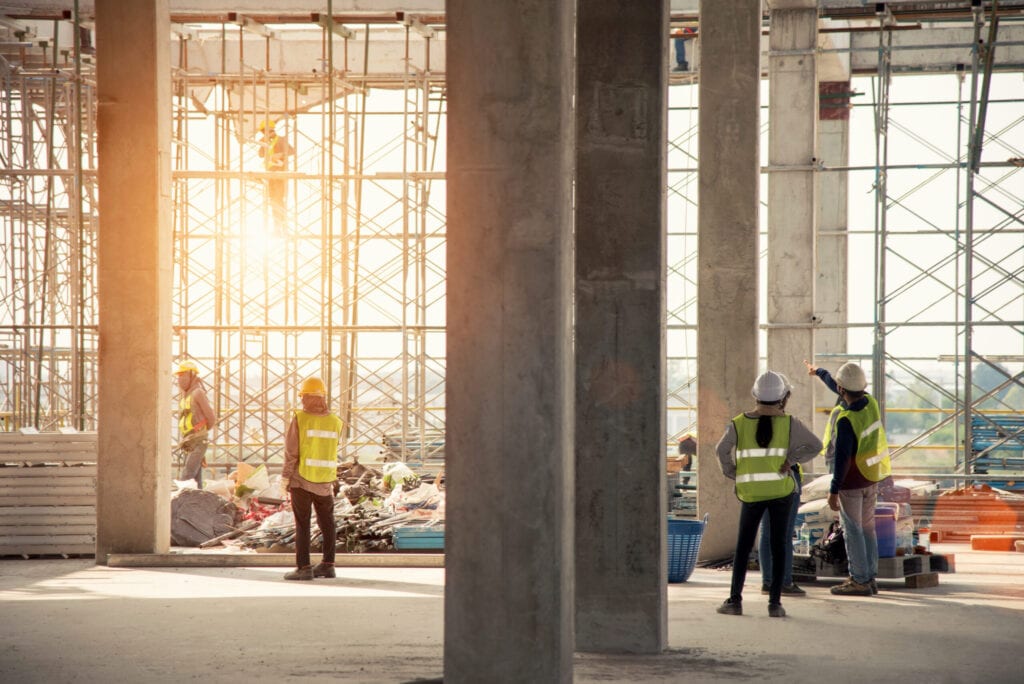 Construction workers in safety vests and helmets at a building site with scaffolding and various materials, under sunlight.