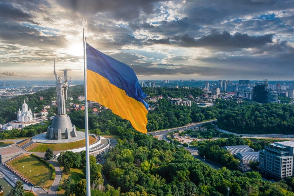 Ukrainian flag waving in front of the Motherland Monument in Kyiv, with a cityscape and dramatic sky in the background.