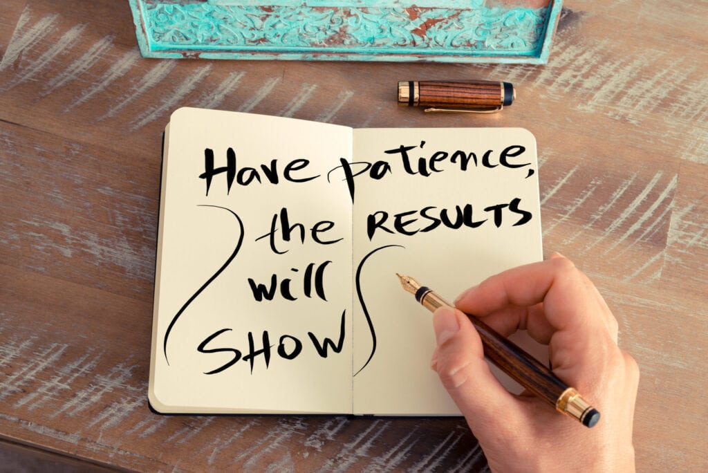 A hand holding a pen next to an open notebook with the words "Have patience, the results will show" written on the pages. An ornate box and fountain pen cap lie on the table.