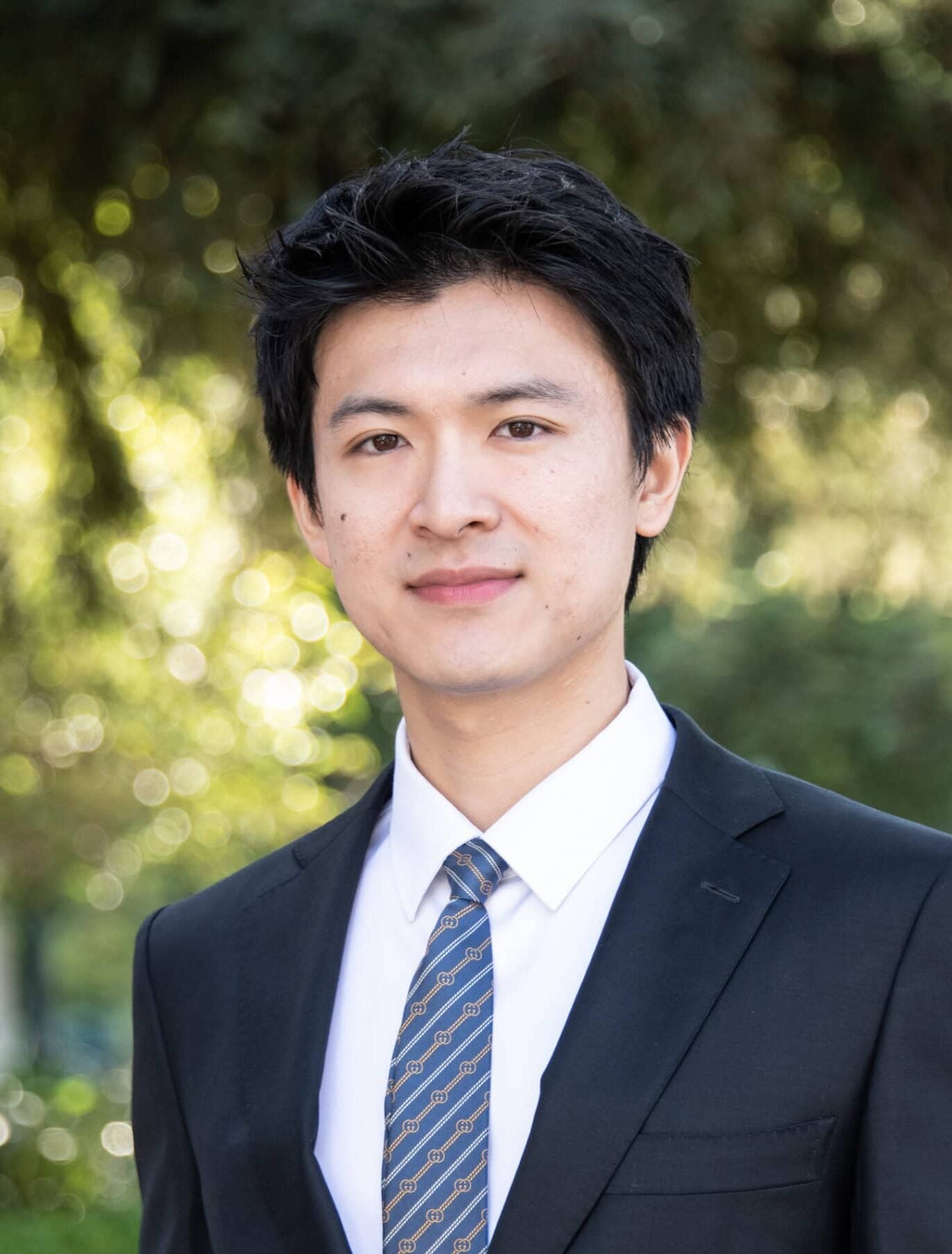 A man in a black suit and patterned tie stands outdoors in front of blurred green foliage, looking at the camera.