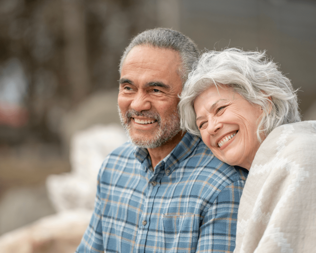 An older couple smiles, with the woman leaning her head on the man's shoulder. They are standing outdoors and wearing casual clothing.