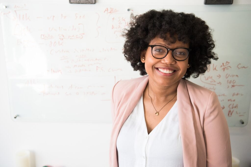 A woman with curly hair and glasses, wearing a white blouse and pink blazer, stands in front of a whiteboard covered in notes and diagrams, smiling at the camera.