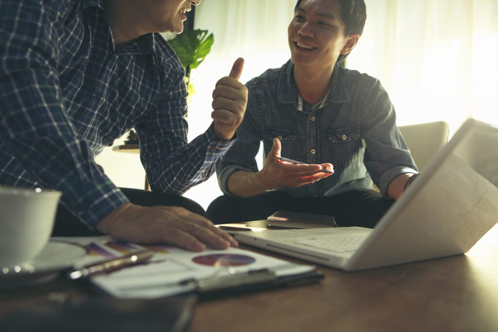 Two people are sitting at a table with a laptop and documents, engaging in a conversation. One person is giving a thumbs-up sign, and both appear to be in a positive mood.