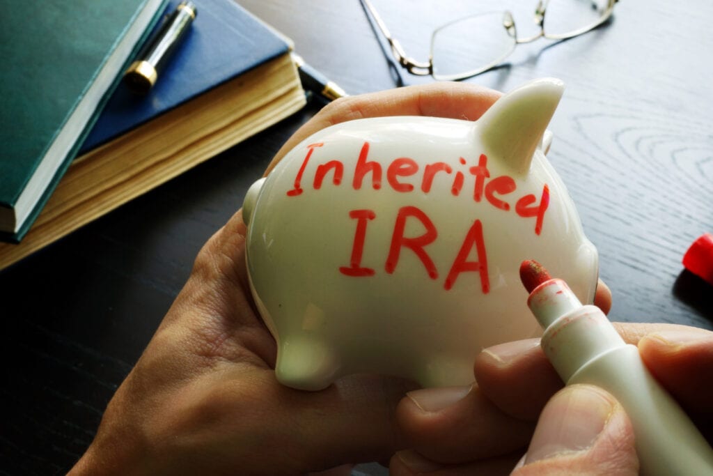 Hands holding a piggy bank with "Inherited IRA" written in red marker. Books, glasses, and a red marker cap are on the black wooden table.