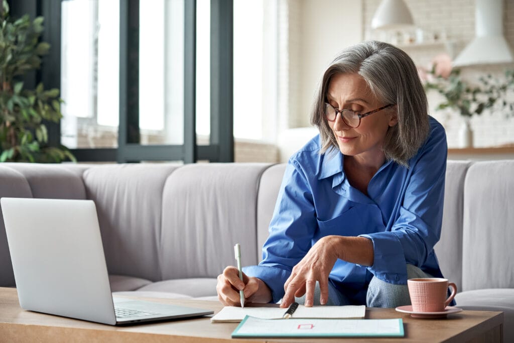 An older woman with gray hair and glasses sits at a table, writing in a notebook while looking at a laptop. A cup is placed beside her on the table.