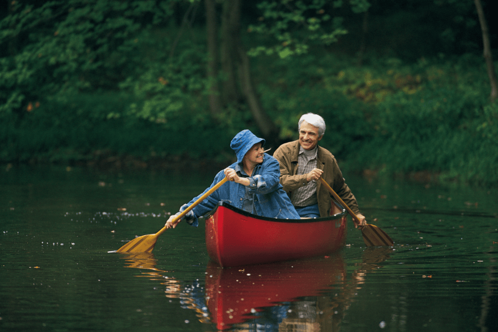 An elderly couple paddles a red canoe on a calm forest lake, surrounded by lush greenery.