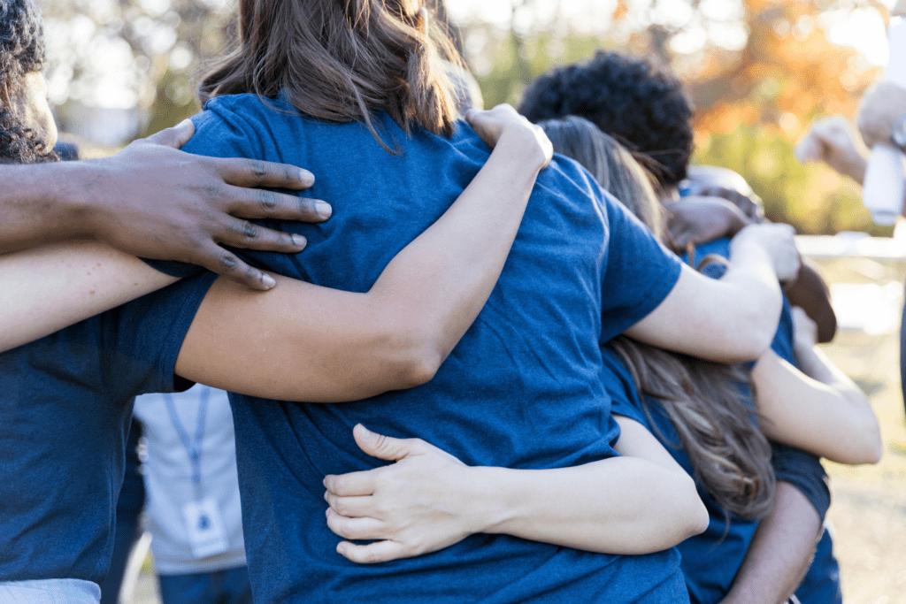 A group of people wearing blue shirts stand in a close huddle, with their arms around each other's shoulders, outdoors.