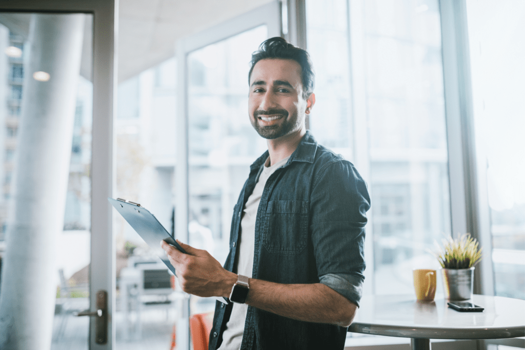A man with a beard, wearing a denim shirt, stands indoors by a table, holding a clipboard and smiling. The background includes large windows and a potted plant.