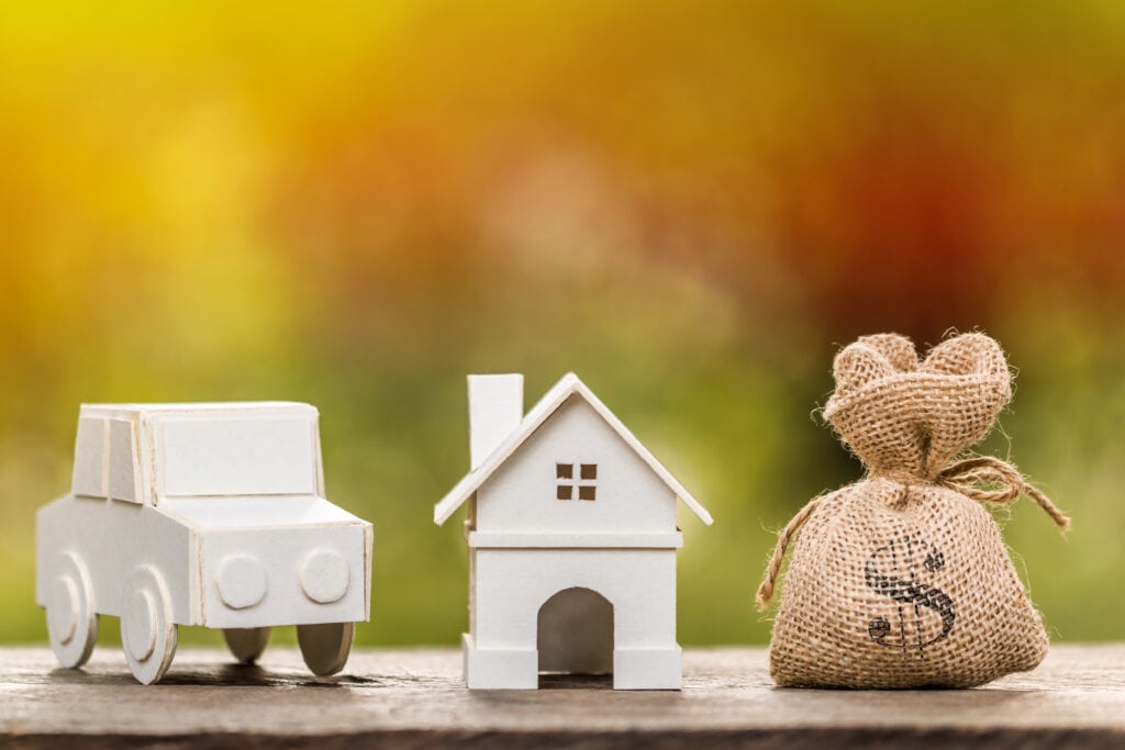 A white model car and a white model house are placed next to a burlap sack with a dollar symbol on it, set against a blurred background.