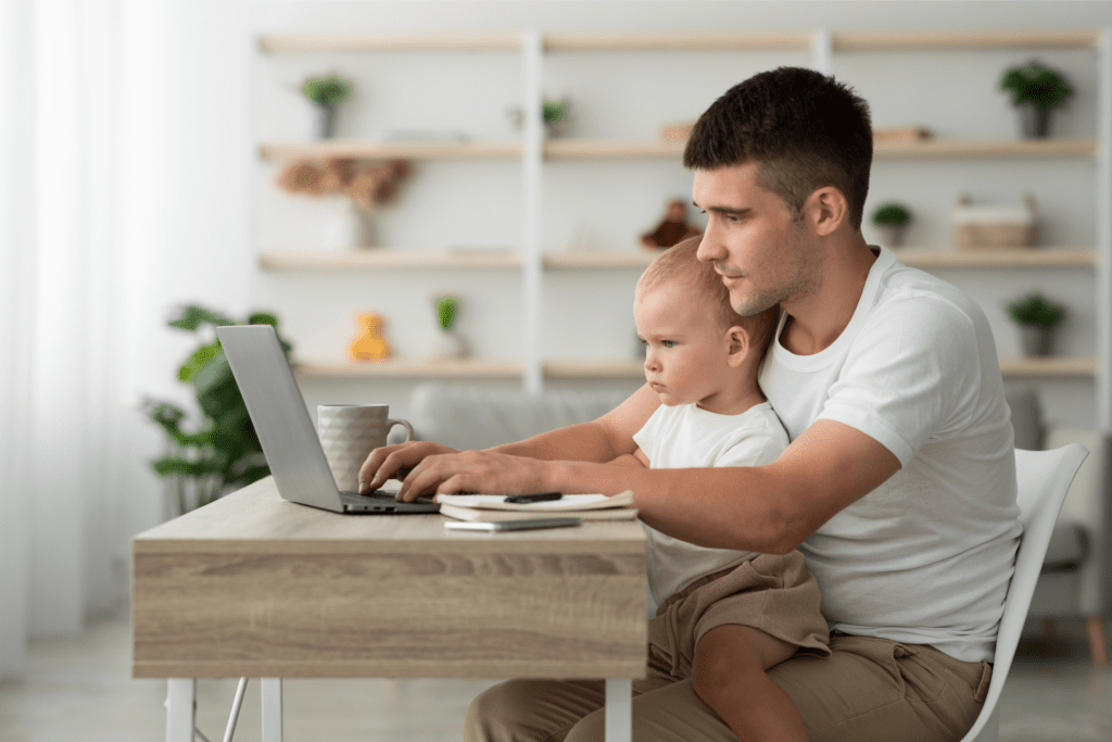 A man is sitting at a desk using a laptop with a child on his lap. There is a notebook and a coffee mug on the desk. The background shows shelves with various items.