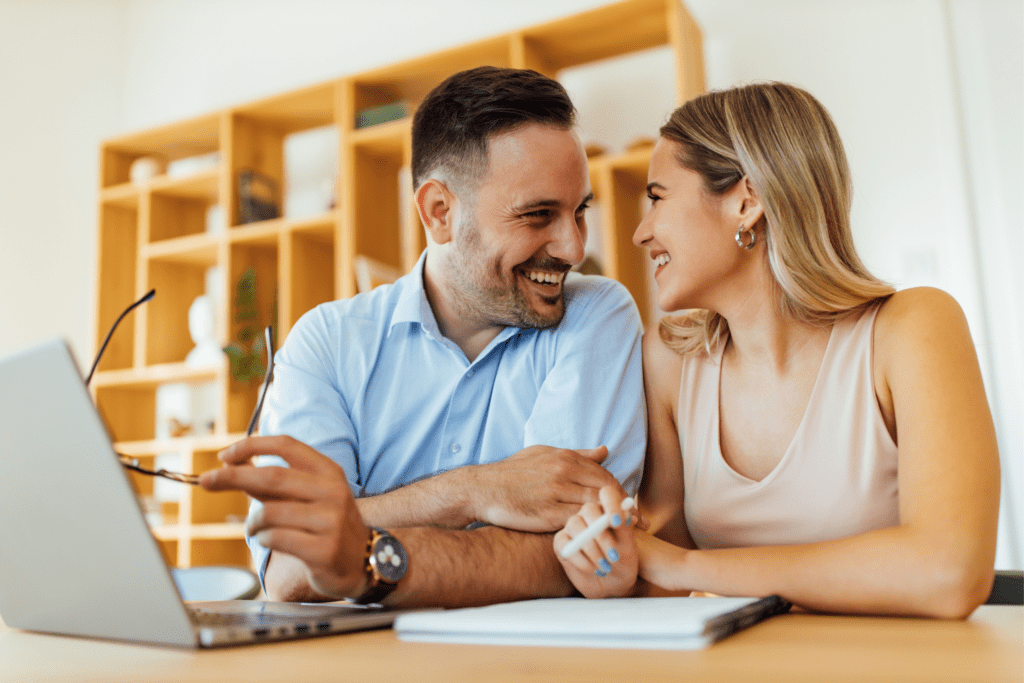 A smiling man and woman sit at a table with a laptop and notebook, looking at each other. The man holds eyeglasses, and there is a wooden shelf in the background.