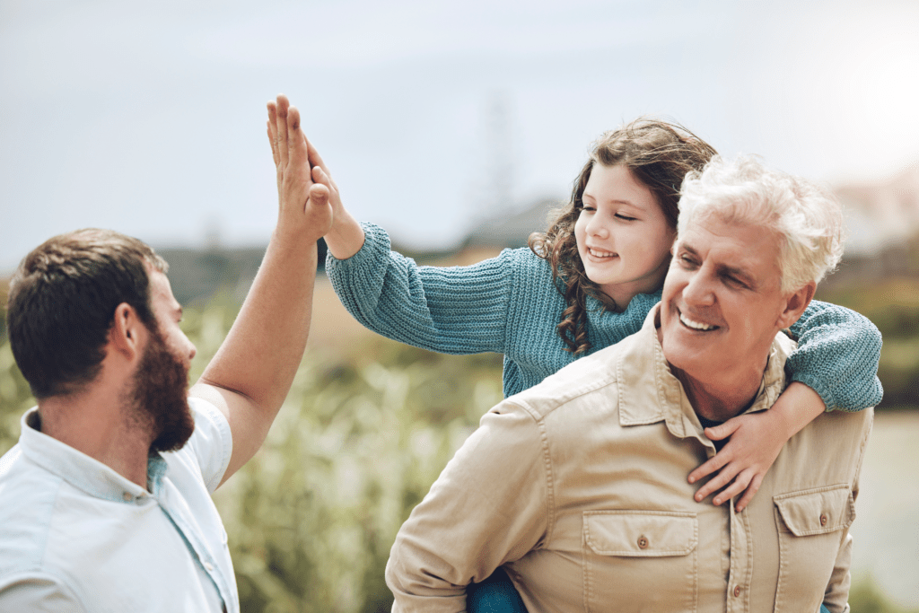 A man holds a child on his back while another man high-fives the child outdoors.