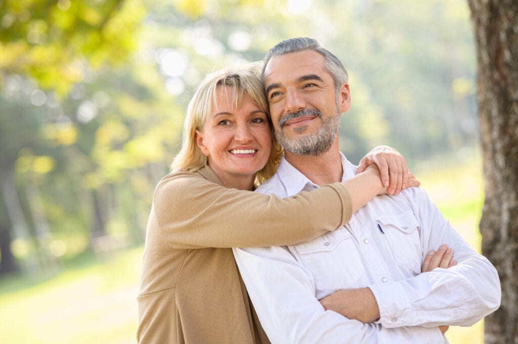 A woman with blond hair hugs a smiling man with graying hair from behind while outdoors in a park.
