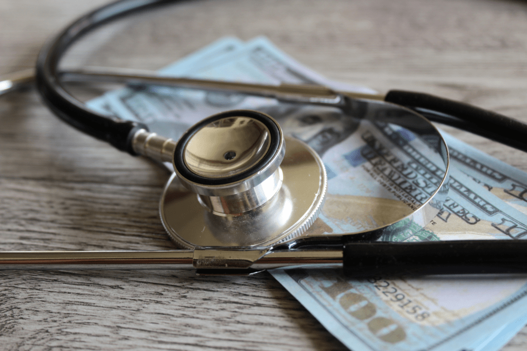 A stethoscope rests on top of several hundred-dollar bills placed on a wooden surface.