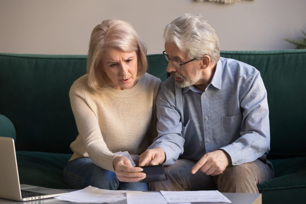 An elderly couple sits on a couch, looking concernedly at a smartphone and documents on the table in front of them, next to a laptop. They seem to be discussing something important.