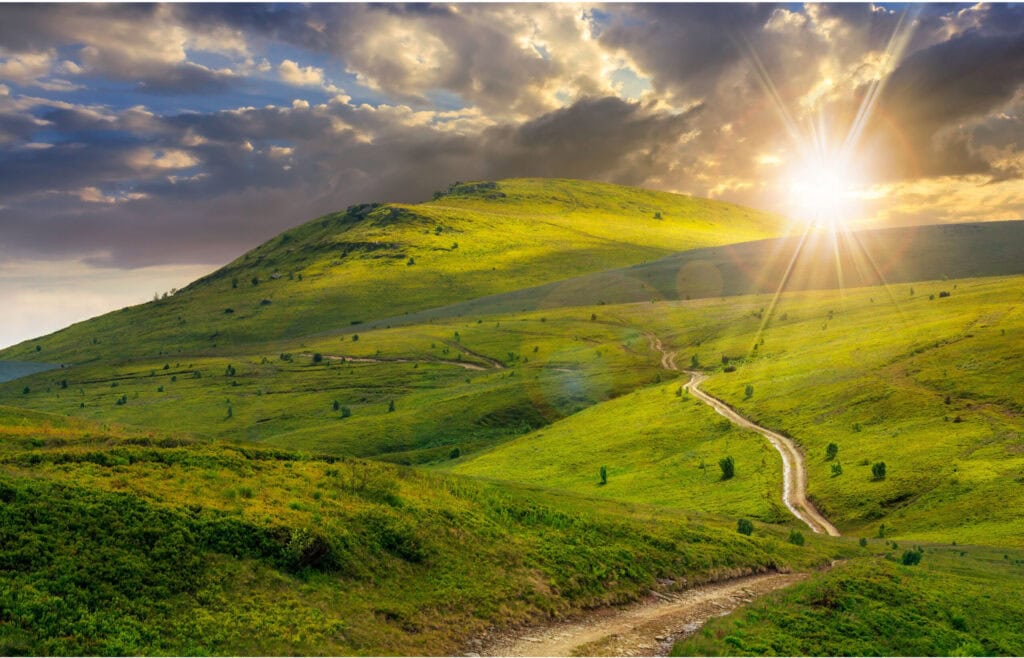 A winding dirt path leads through rolling green hills toward a bright setting sun, with a dramatic sky filled with scattered dark clouds.