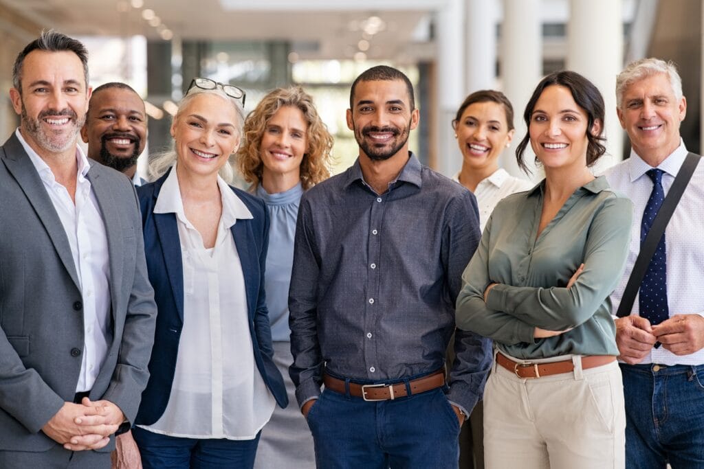 A diverse group of eight smiling professionals, both men and women, standing indoors in a well-lit office environment.