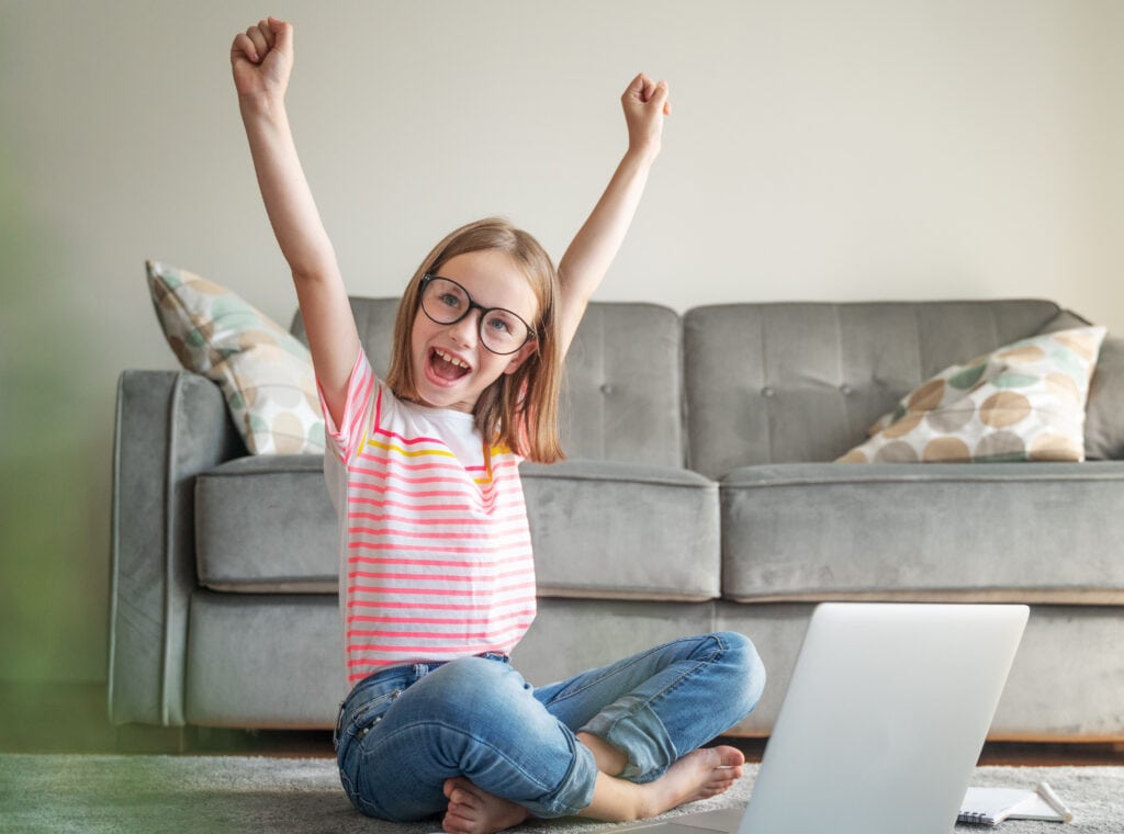 A child with glasses sits cross-legged on the floor, raising both arms in excitement, with a laptop in front and a sofa in the background.