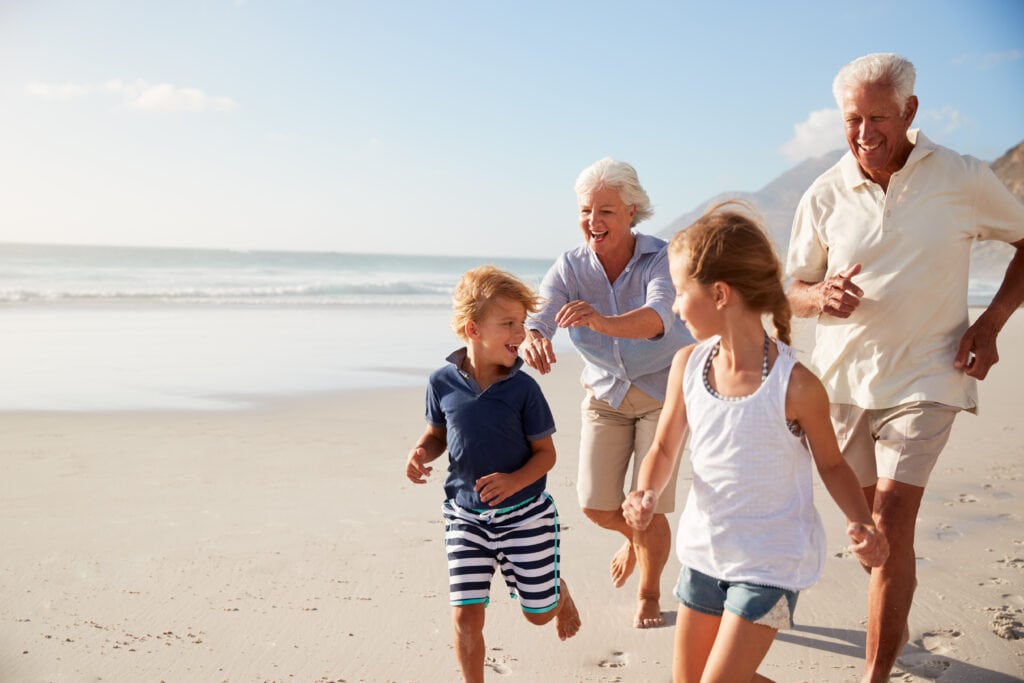 Two adults and two children run joyfully together on a sunny beach with waves in the background. The adults appear older, possibly grandparents, and the children are a boy and a girl.