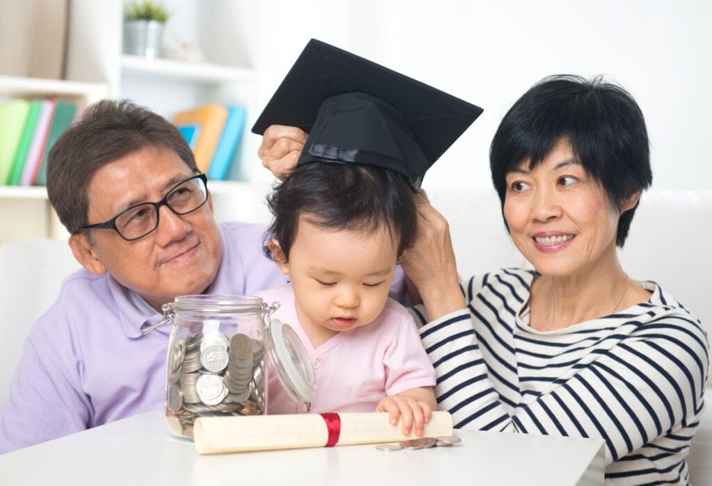 An elderly couple with glasses smiles at a toddler wearing a graduation cap. The child is near a jar of coins and a rolled-up diploma tied with a red ribbon.