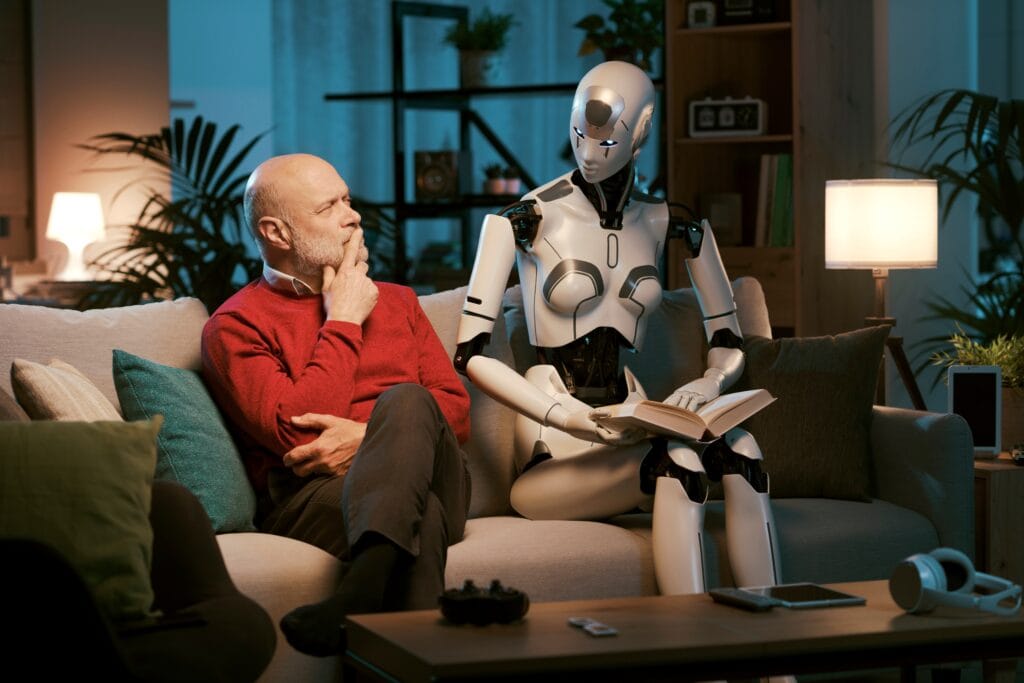 An elderly man and a humanoid robot sit on a couch; the robot is holding a book while the man looks at it thoughtfully. The setting appears to be a modern living room.