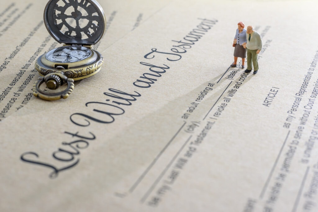 A pocket watch and small figurines of two older people stand on a document titled "Last Will and Testament".