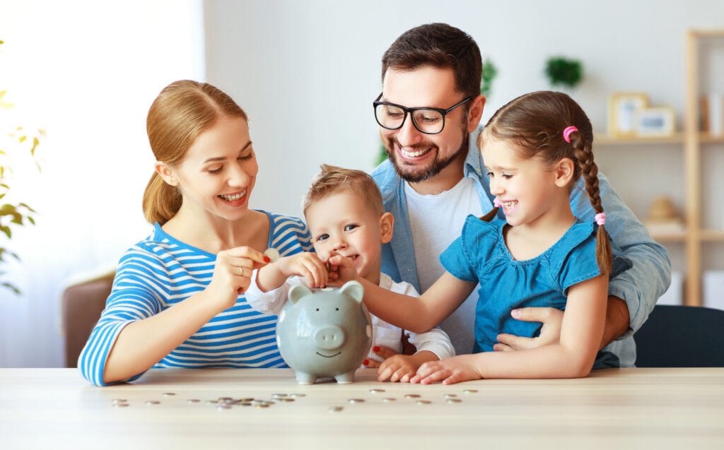 A smiling family of four, including two children, are sitting at a table placing coins into a gray piggy bank.
